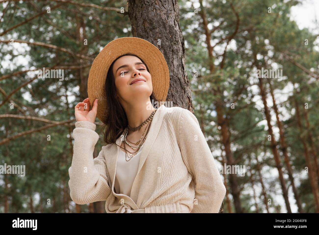 Fröhliche junge Frau, die Strohhut anpasst und im Wald lächelt Stockfoto