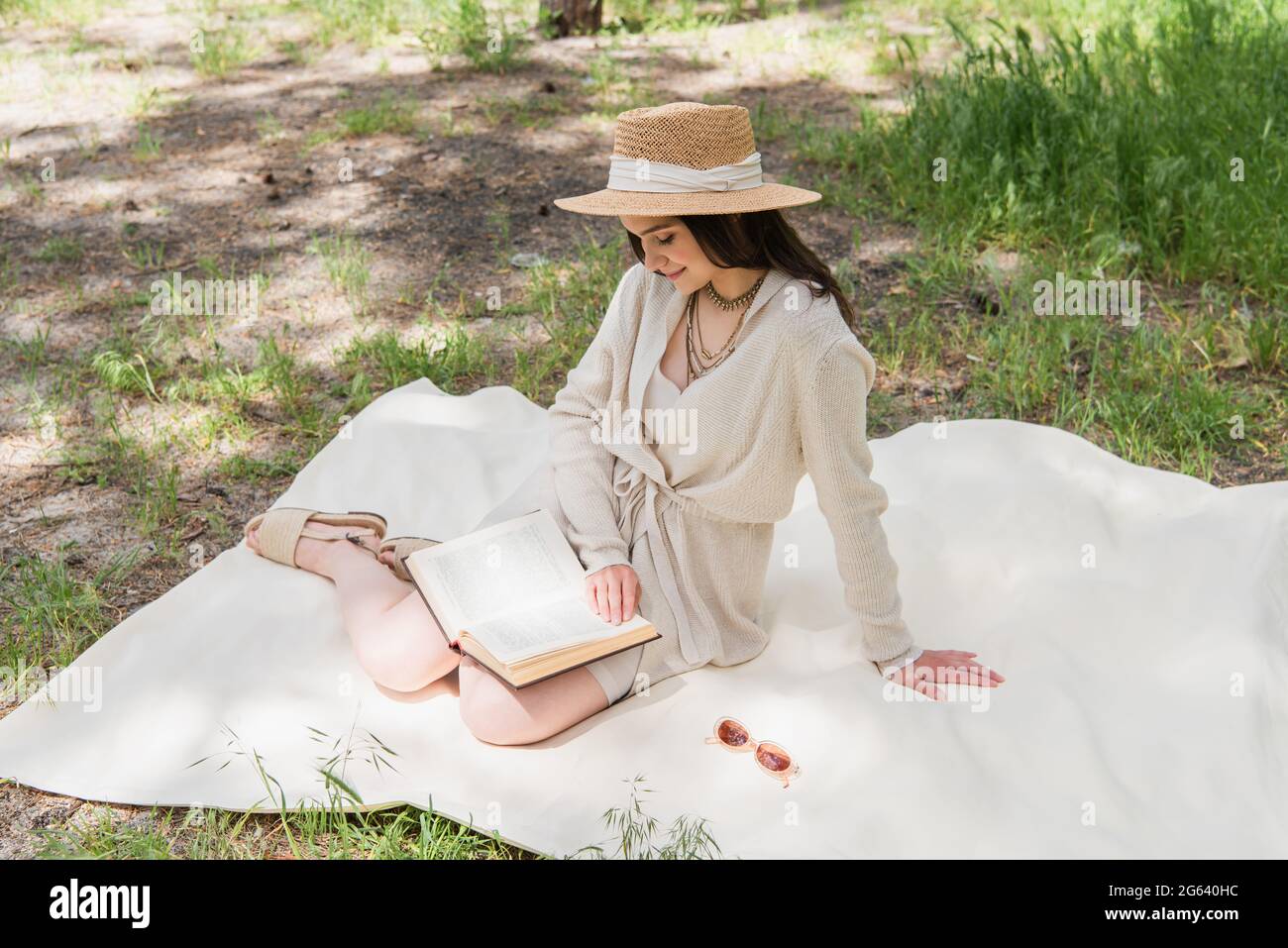 Lächelnde junge Frau im Strohhut liest Buch im Wald Stockfoto