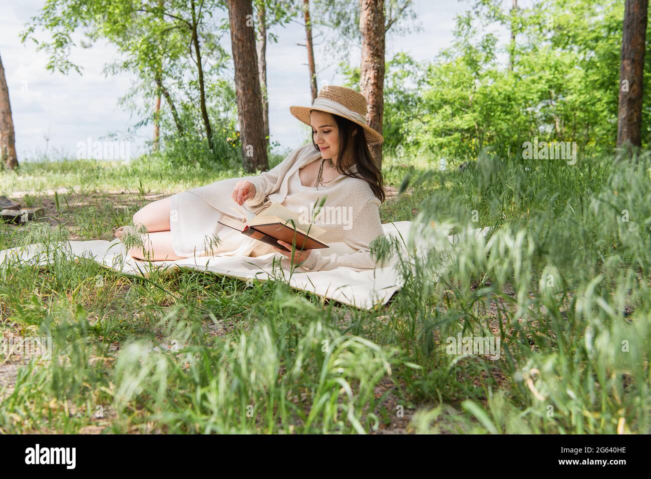 Fröhliche junge Frau im Strohhut, die Buch im Wald liest Stockfoto