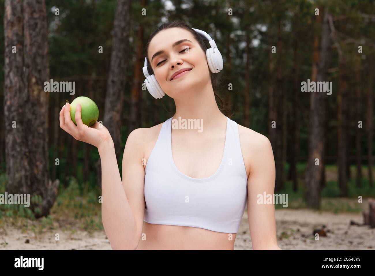 Fröhliche Frau in Kopfhörern, die Musik hört, während sie den Apfel draußen hält Stockfoto