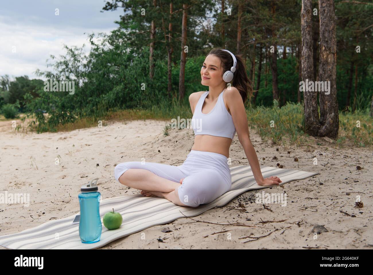 Lächelnde Frau in Kopfhörern, die Musik hört, während sie mit gekreuzten Beinen auf einer Yogamatte sitzt Stockfoto