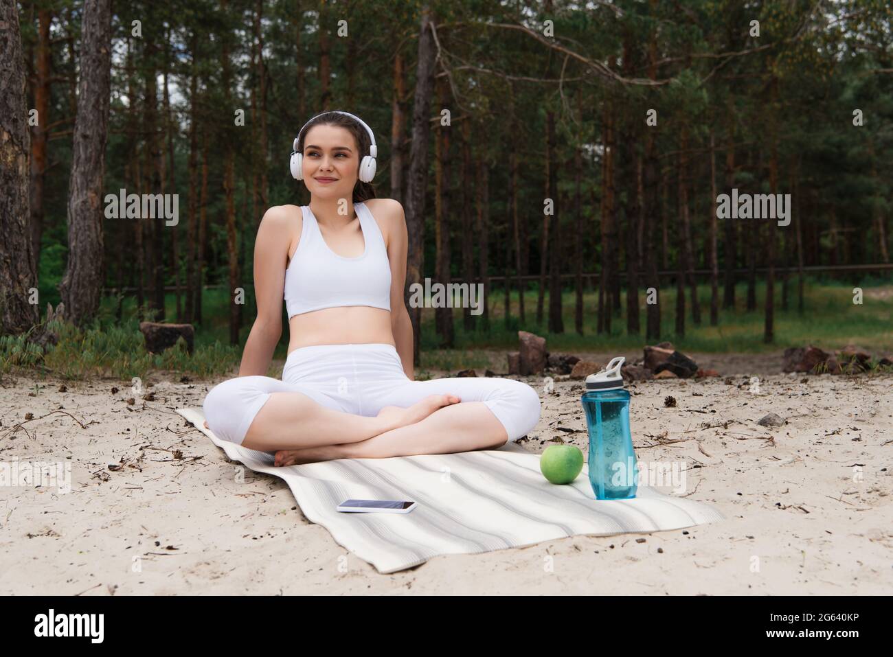Fröhliche Frau in Kopfhörern, die Musik hört, während sie mit gekreuzten Beinen auf einer Yogamatte sitzt Stockfoto