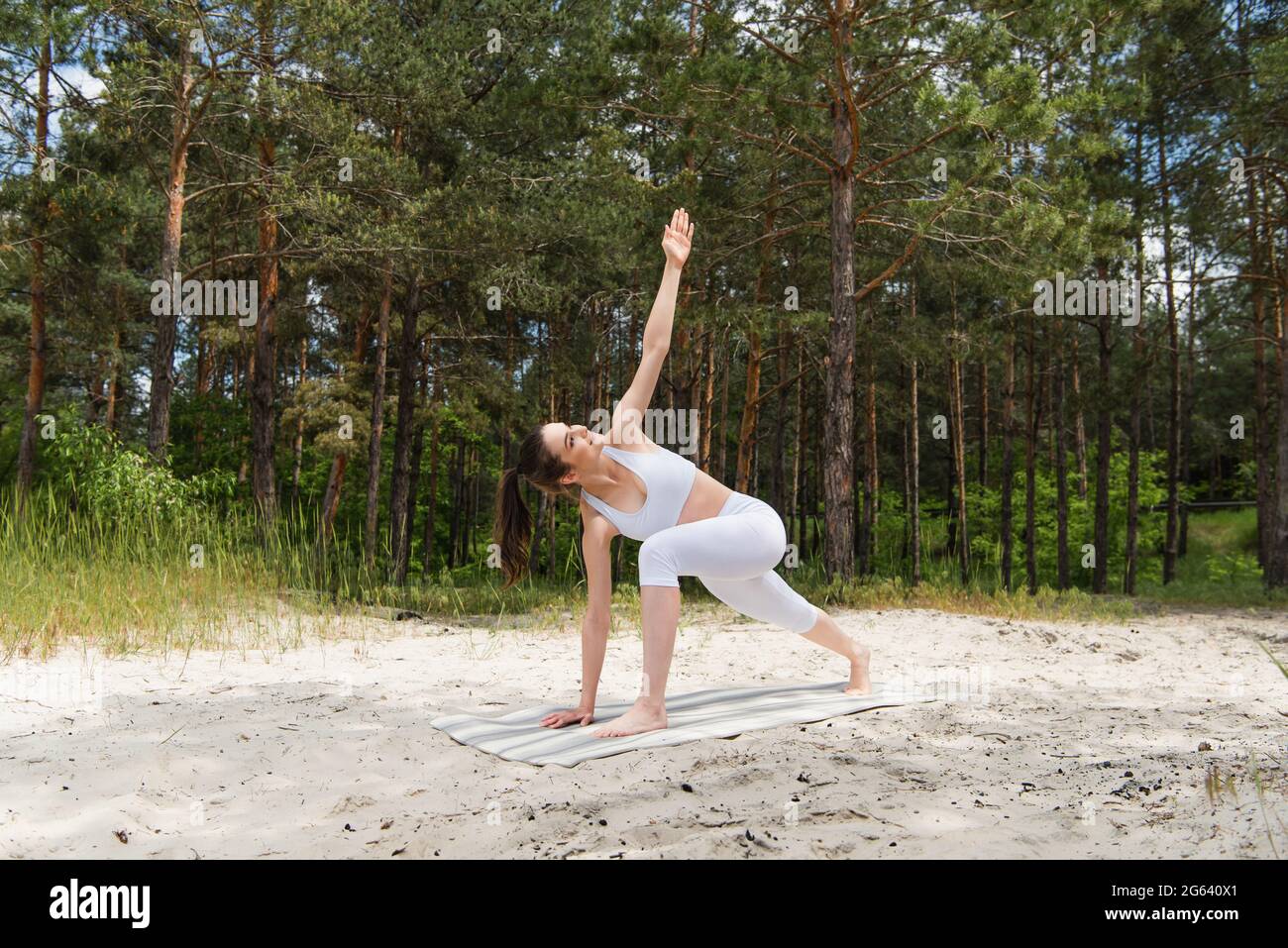 In voller Länge eine glückliche junge Frau, die im Wald einen Ausfallschritt auf der Yogamatte macht Stockfoto