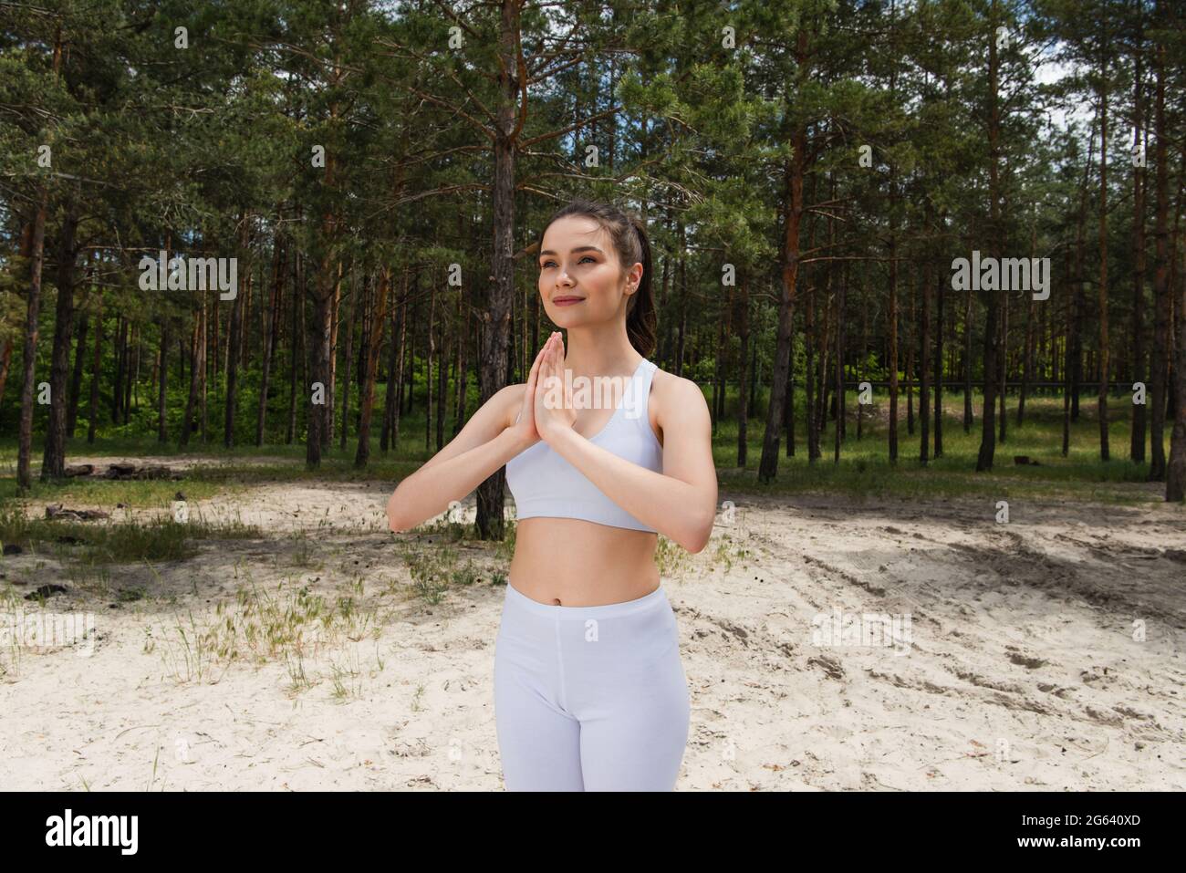 Lächelnde junge Frau im Ernteguttop und Leggings, die mit betenden Händen im Wald stehen Stockfoto