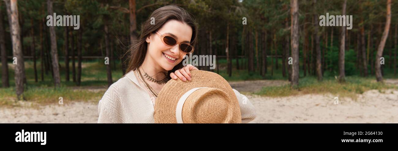 Fröhliche junge Frau in Sonnenbrille mit Strohhut in der Nähe des Waldes, Banner Stockfoto