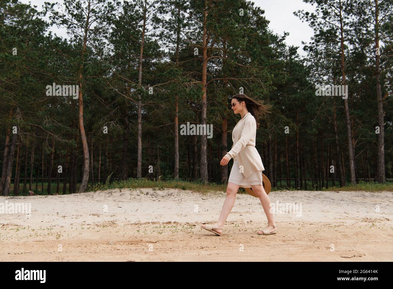 Die ganze Länge der fröhlichen jungen Frau in Sonnenbrillen mit Strohhut und Spaziergang in der Nähe des Waldes Stockfoto