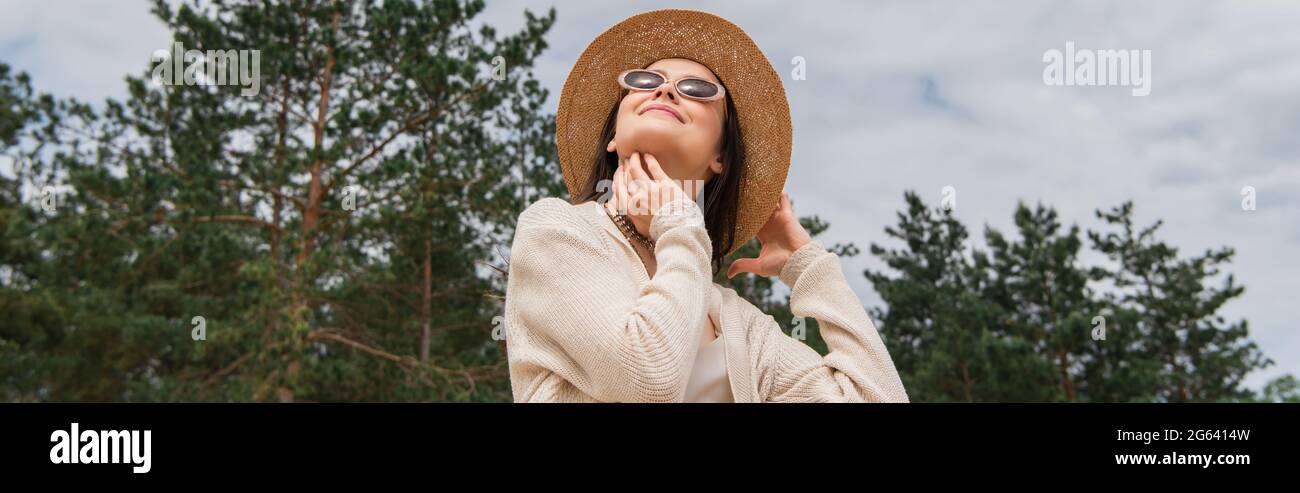 Low-Angle-Ansicht der fröhlichen jungen Frau in Sonnenbrille und Strohhut in der Nähe von Wald, Banner Stockfoto