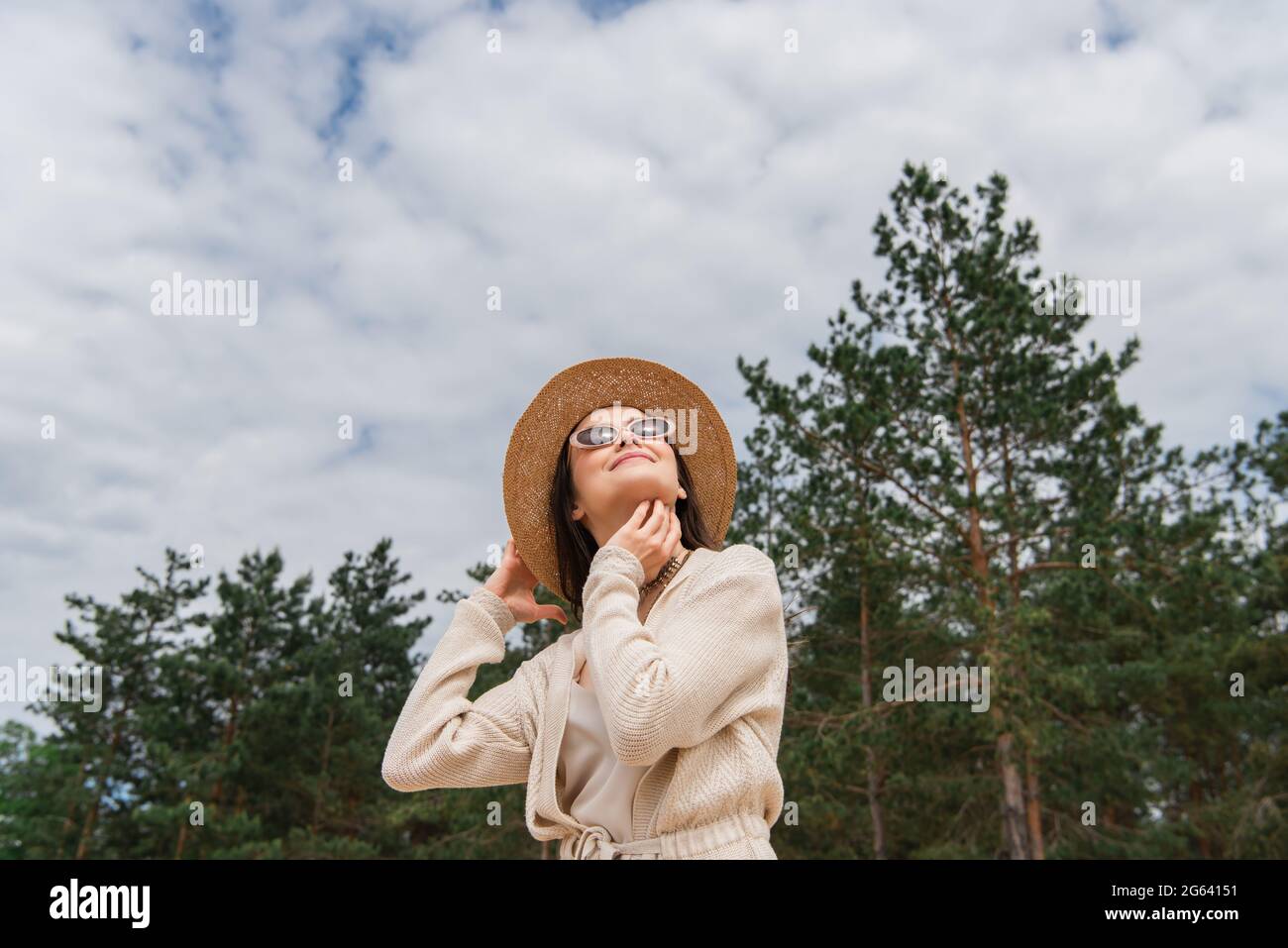 Blick aus der unteren Perspektive auf eine fröhliche junge Frau mit Sonnenbrille und Strohhut, die in der Nähe des Waldes aufschaut Stockfoto