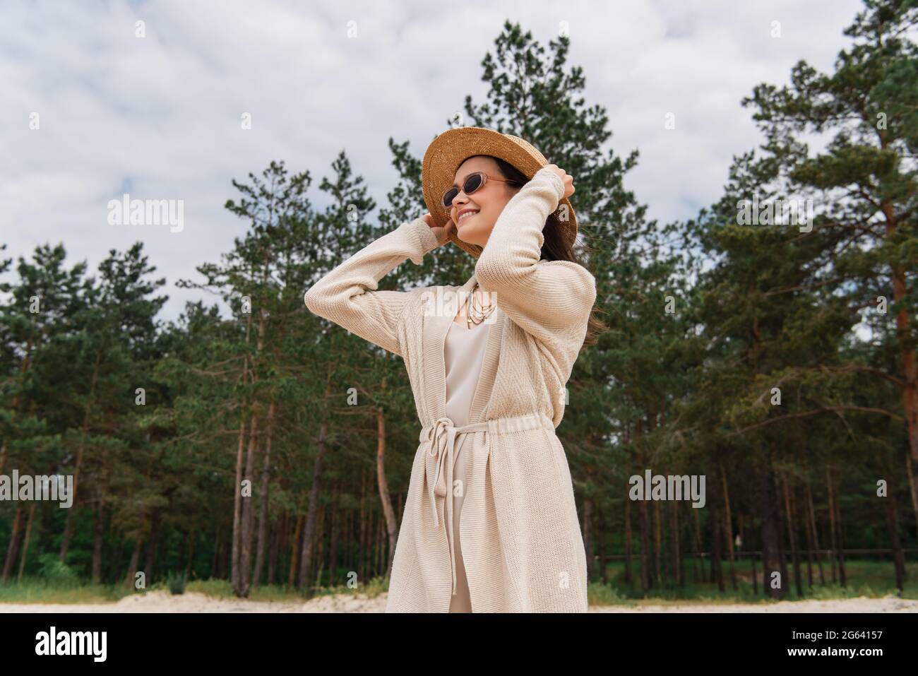Fröhliche junge Frau in Sonnenbrillen Anpassung Strohhut in der Nähe des Waldes Stockfoto