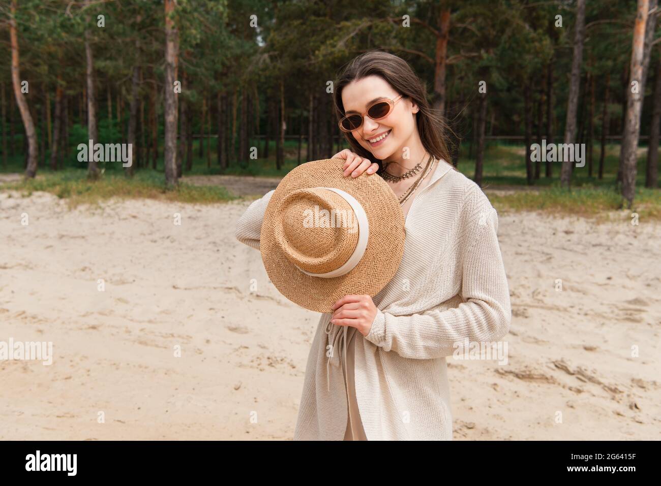 Fröhliche junge Frau in Sonnenbrille mit Strohhut in der Nähe des grünen Waldes Stockfoto