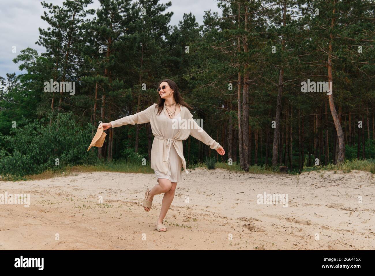 Die ganze Länge der positiven jungen Frau in einer Sonnenbrille hält Strohhut und geht in der Nähe des Waldes Stockfoto