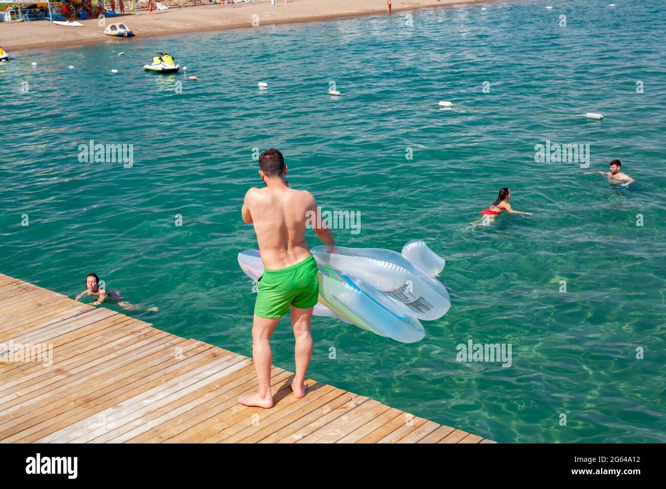 Antalya, Türkei-28. Juni 2021: Mann mit aufblasbarer Luftmatratze auf Holzdock, während andere Strandbesucher im Sommer in Antalya im Meer schwimmen. Stockfoto