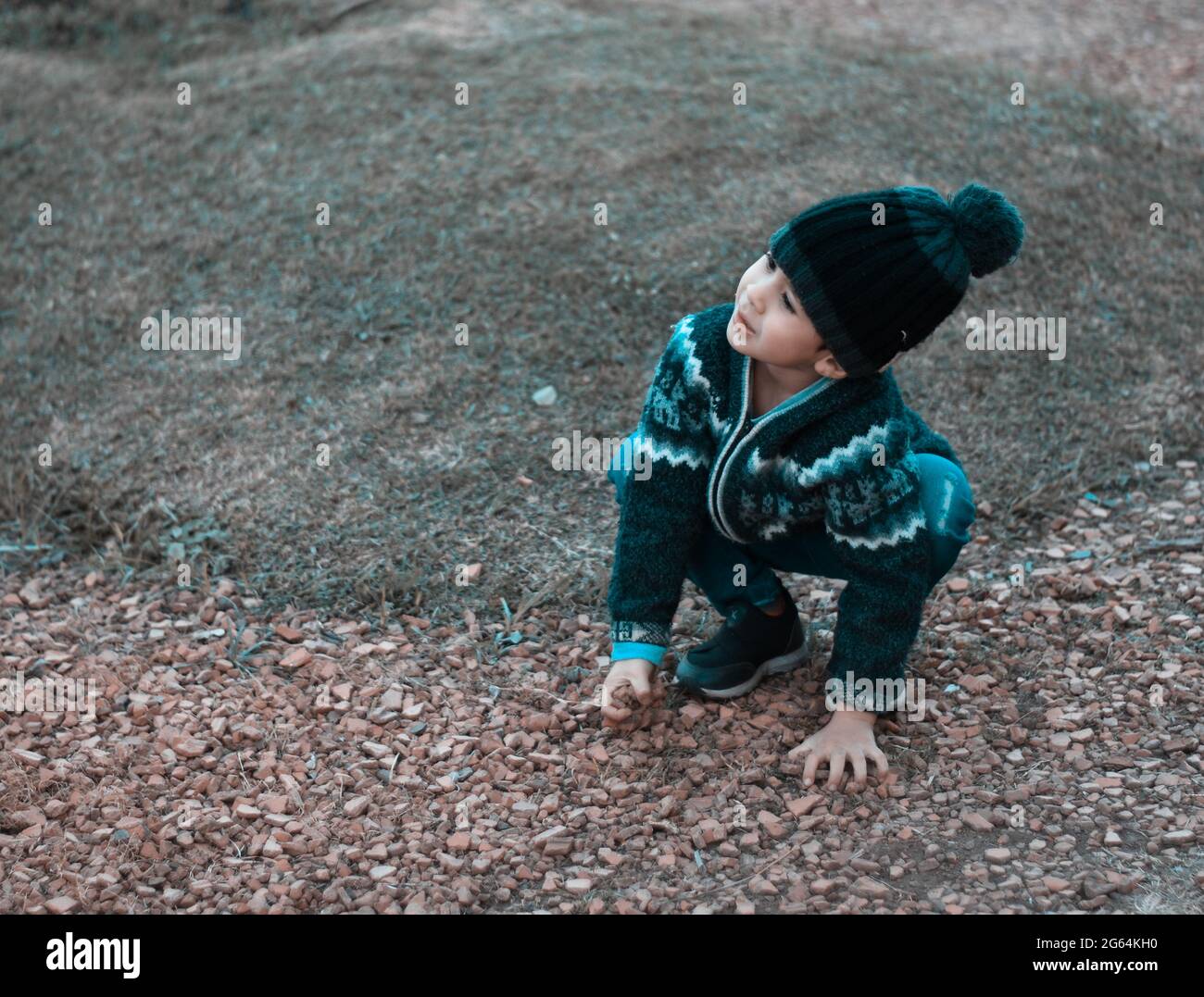 Kind hockend unten spielend mit Kieselsteinen im Park und schaut oben im Winter in einem Wollhut, waagerecht Stockfoto
