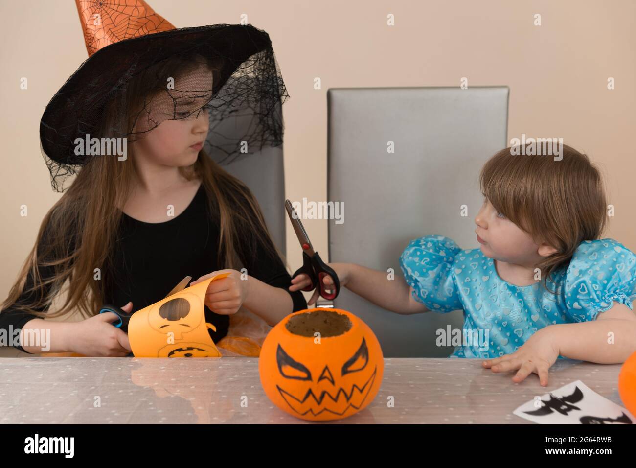 Zwei Mädchen Schwestern in Kostümen einer Hexe und Schneewittchen, die an einem Tisch sitzen, schneiden eine Papiergirlande zur Dekoration für den Halloween-Urlaub aus Stockfoto