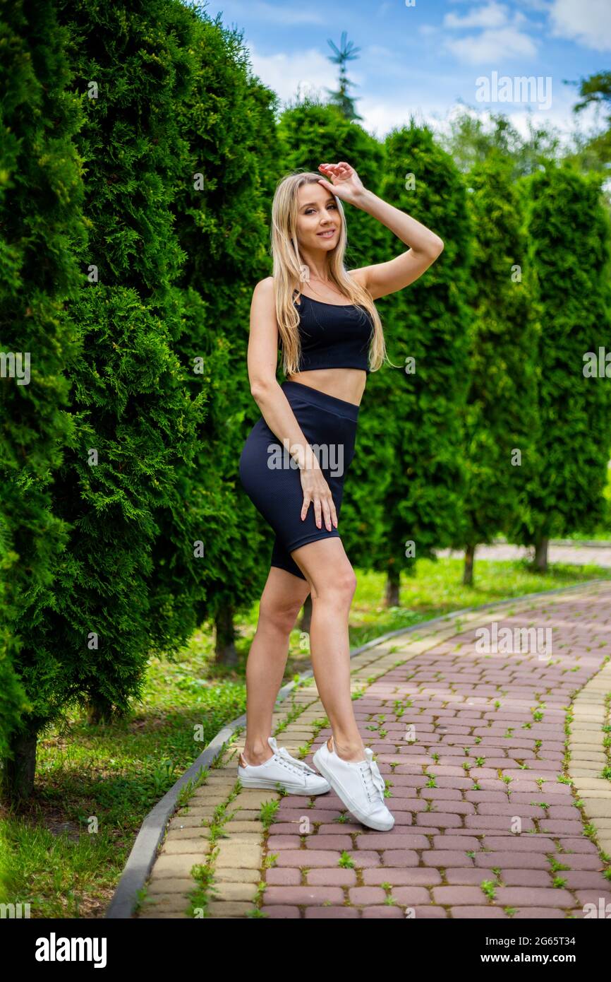 Junge schöne sportliche blonde Frau in einem schwarzen T-Shirt und in schwarzen engen Sport-Shorts in guter Form posiert vor grünen Bäumen und Büschen in nat Stockfoto