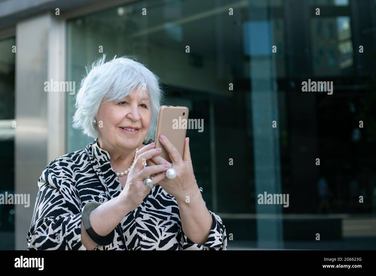Lächelnde grauhaarige ältere Frau mit Mobiltelefon in der Hand auf der Straße Stockfoto
