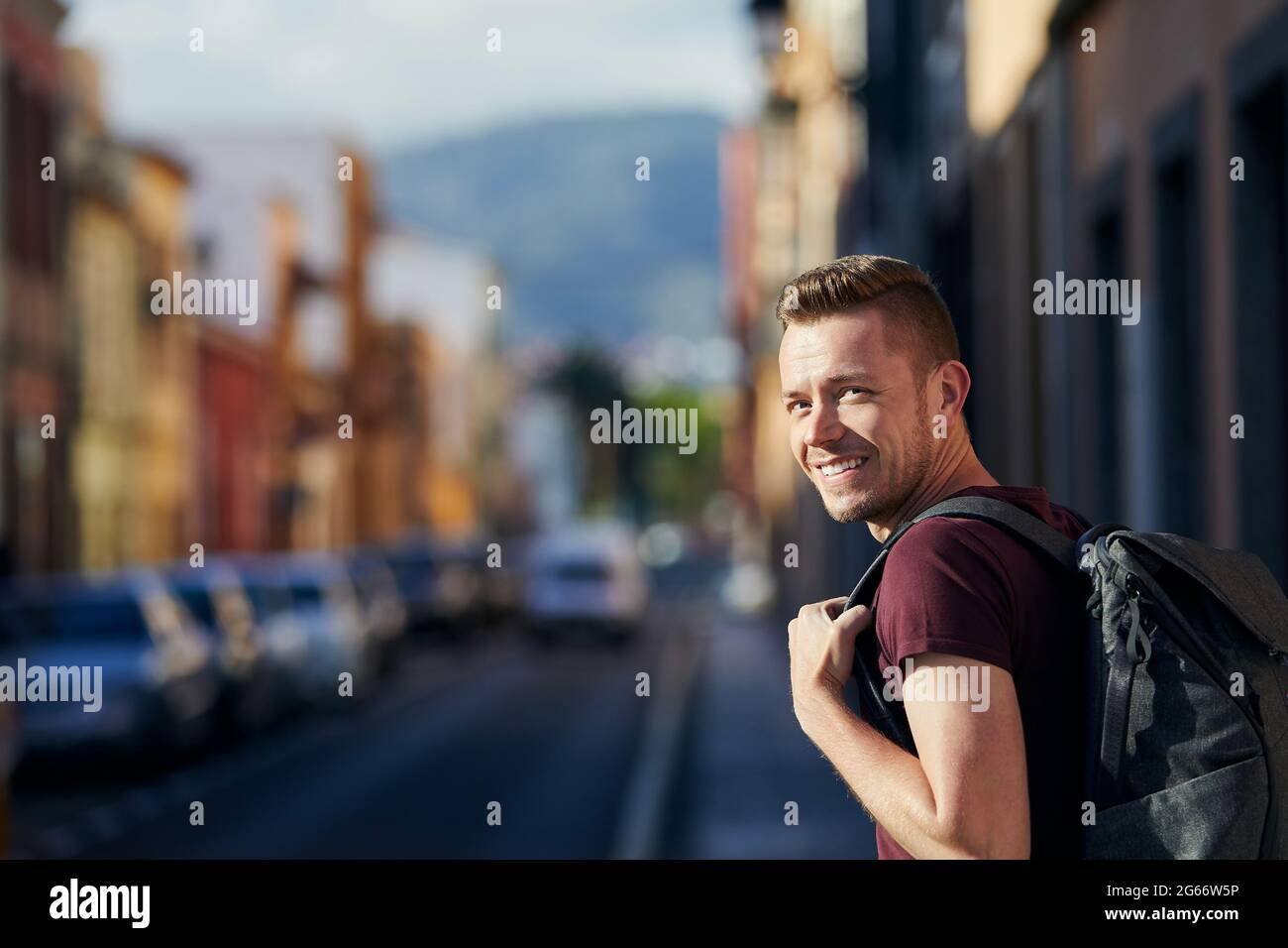 Glücklicher Alleinreisender, der die Kamera anschaut. Lächelnder Mann mit Rucksack auf der Straße der Altstadt. San Cristóbal de La Laguna auf Teneriffa, Kanarische Inseln, Spanien. Stockfoto
