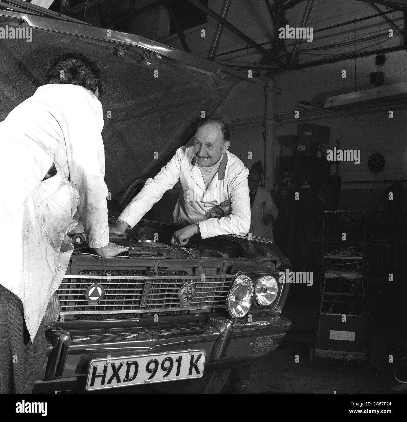 1970s, historisch, in einer Autowerkstatt, zwei Garagenmechaniker, die neben einem Motorwagen mit aufstehender Motorhaube stehen, den Motor des Autos betrachten und versuchen herauszufinden, was das Problem ist, England, Großbritannien. Stockfoto