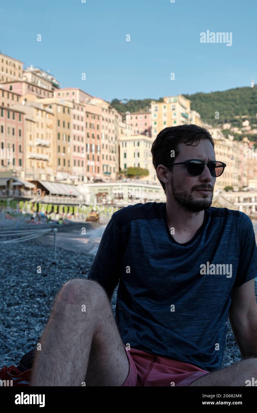 Ein Porträt eines jungen Models mit Sonnenbrillen am Strand von Camogli, Genua, Italien, mit den bunten Häusern dahinter Stockfoto