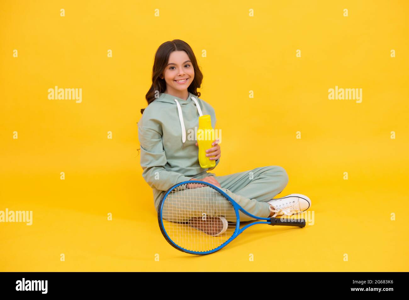Glückliches Kind sitzt in Sportbekleidung mit Tennis-Schläger Wasserflasche auf gelbem Hintergrund, Wasserhaushalt Stockfoto