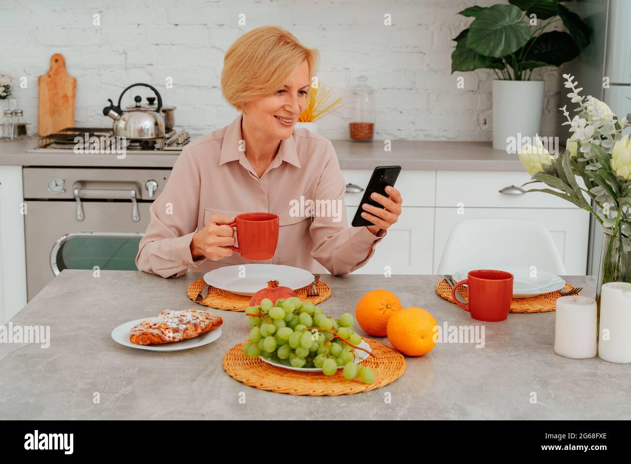 Die Frau frühstückt zu Hause mit Obst, Kuchen und Kaffee und liest Nachrichten vom Smartphone Stockfoto