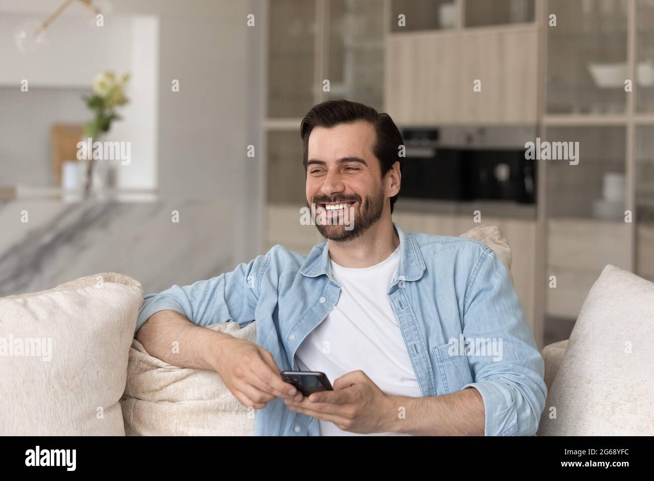 Überglücklich lachender junger Mann lenkte vom Smartphone ab, das auf der Couch saß Stockfoto
