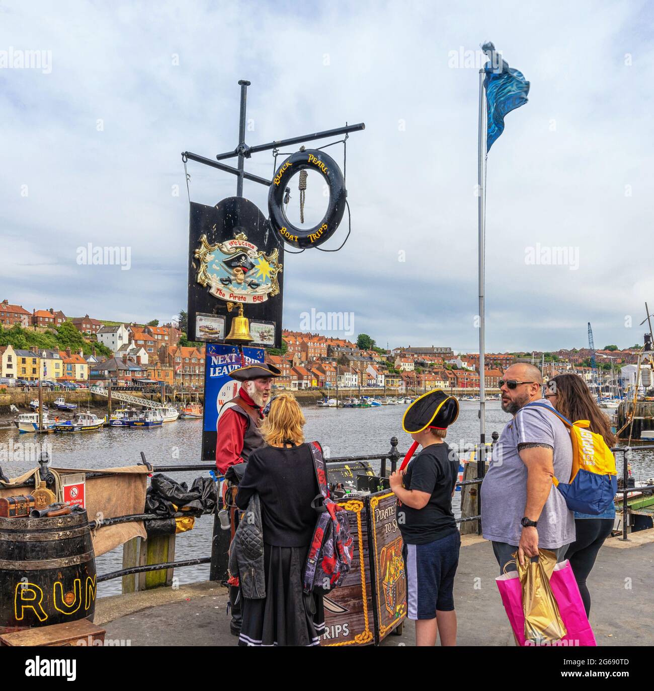 Touristen versammeln sich um einen Stand mit Piraten-Thema und verkaufen Tickets für eine Vergnügungsbootfahrt. Ein Hafen ist dahinter und ein wolkiger Himmel ist oben, Stockfoto