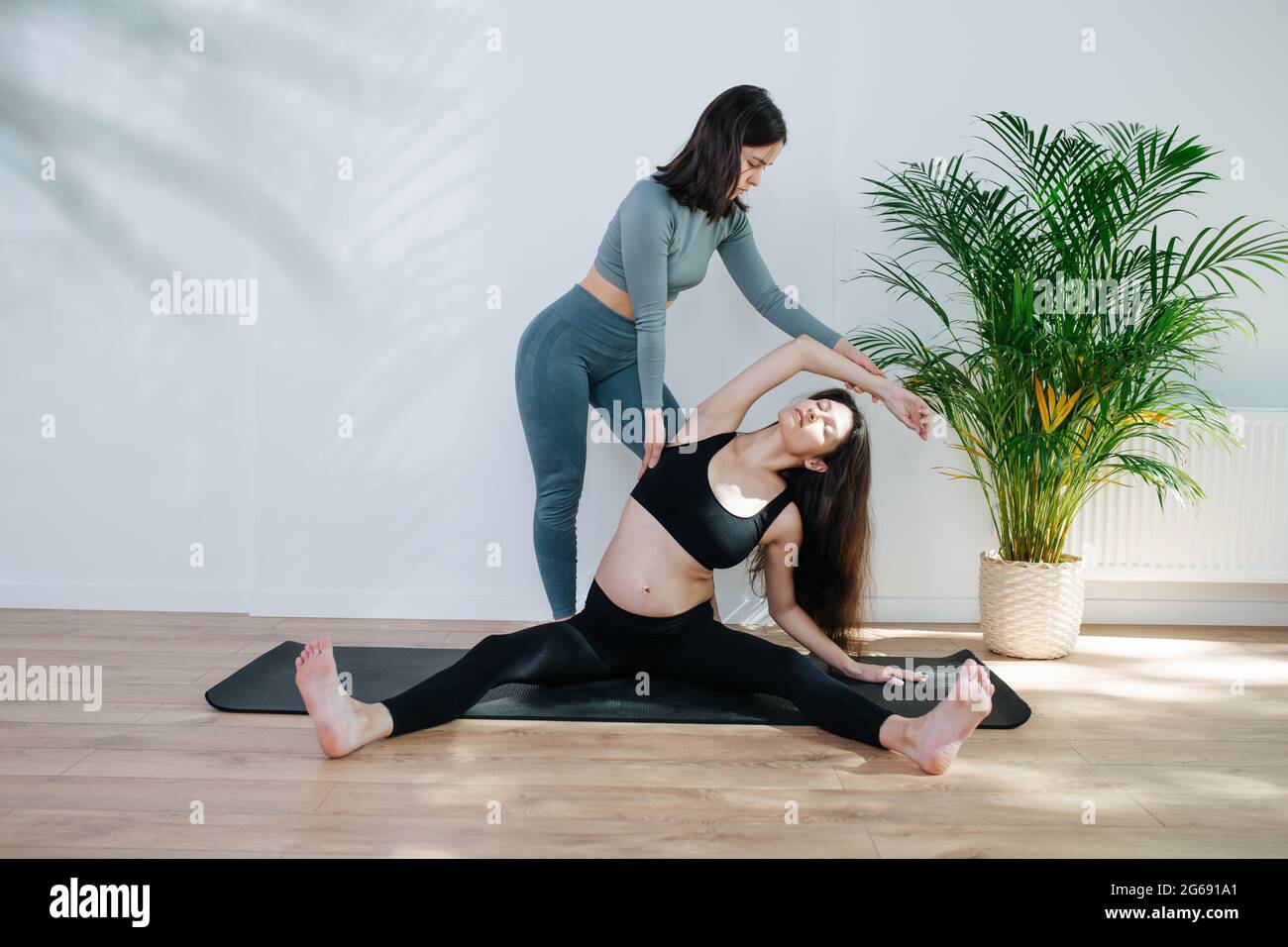 Trainer, der Schwangeren hilft, Yoga zu praktizieren, sie machen tiefe Seitenstrecken. Frau sitzt auf einer Matte mit gespreizten Beinen in einem großen Studio. Sie ist wea Stockfoto