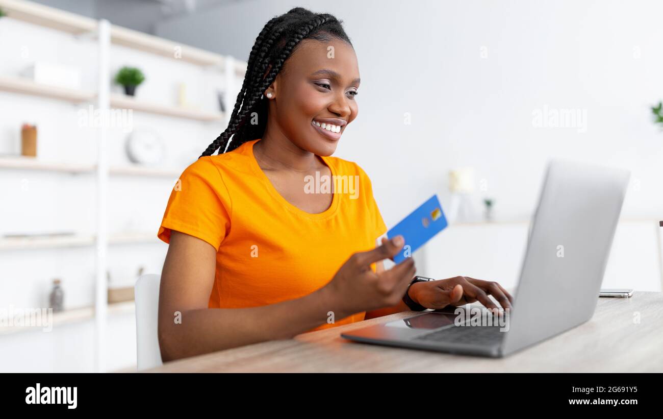 Online-Shopping. Junge schwarze Dame mit Laptop und Kreditkarte sitzt am Tisch im Haus und kauft Waren im Webshop Stockfoto