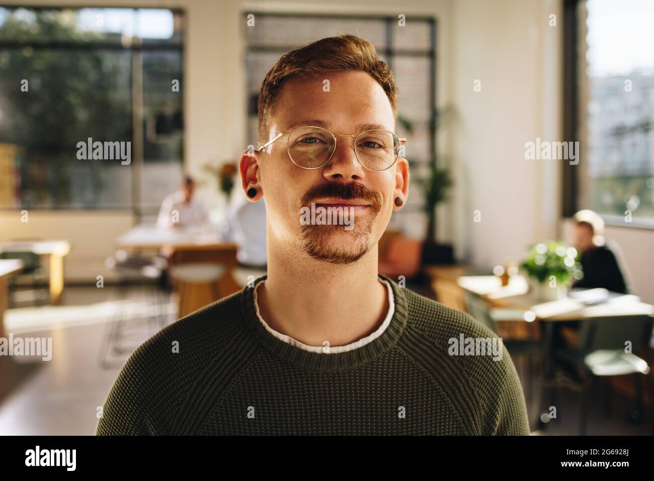 Geschäftsmann lächelt, während er im kooperativen Bereich mit Leuten steht, die hinten arbeiten. Glücklicher Mann, der im Amt steht und Kollegen arbeiten. Stockfoto