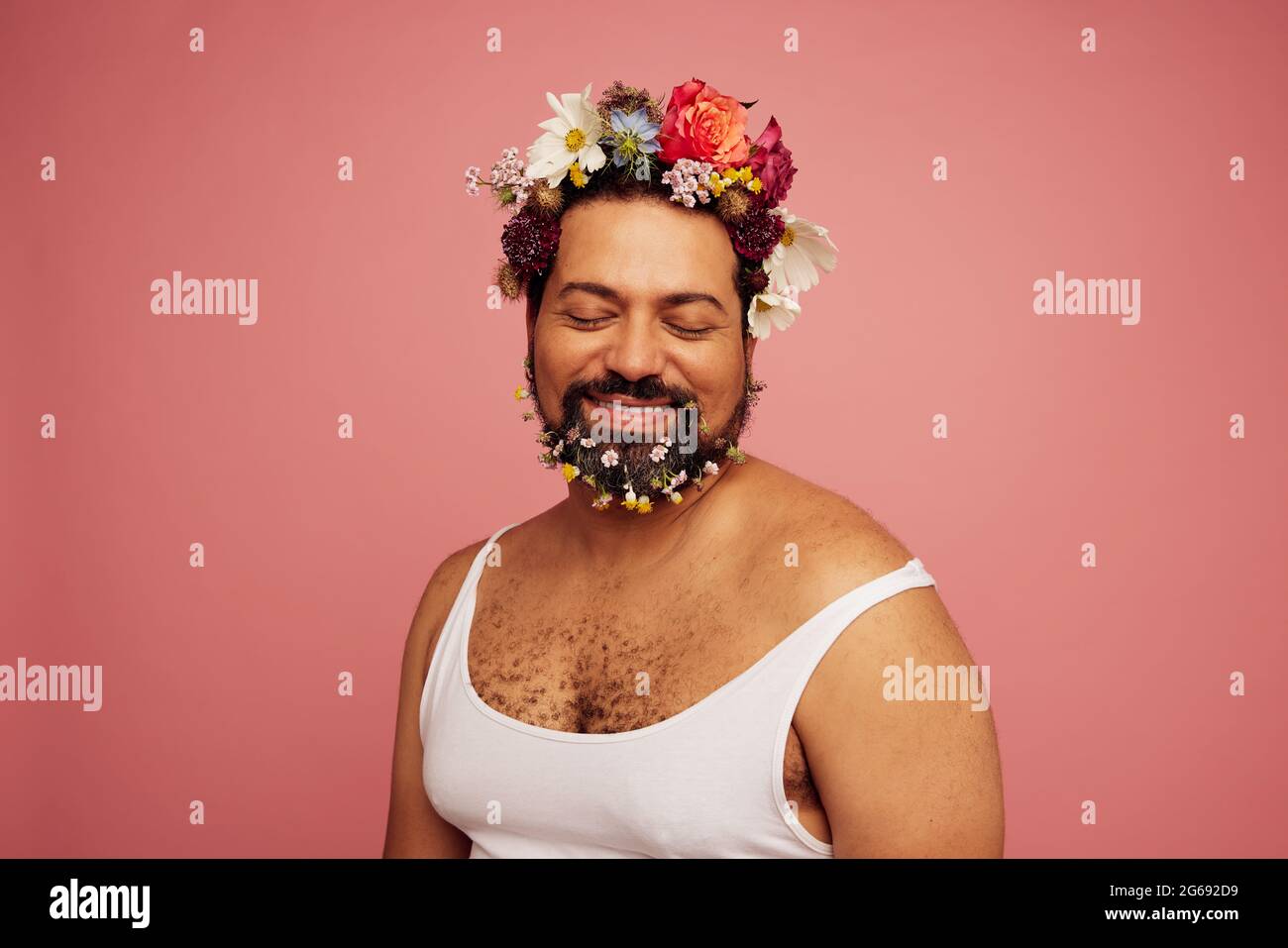 Nicht binäre Person trägt Tank Top posiert auf rosa Hintergrund. Genderqueer trägt Blumen auf Kopf und Bart. Stockfoto