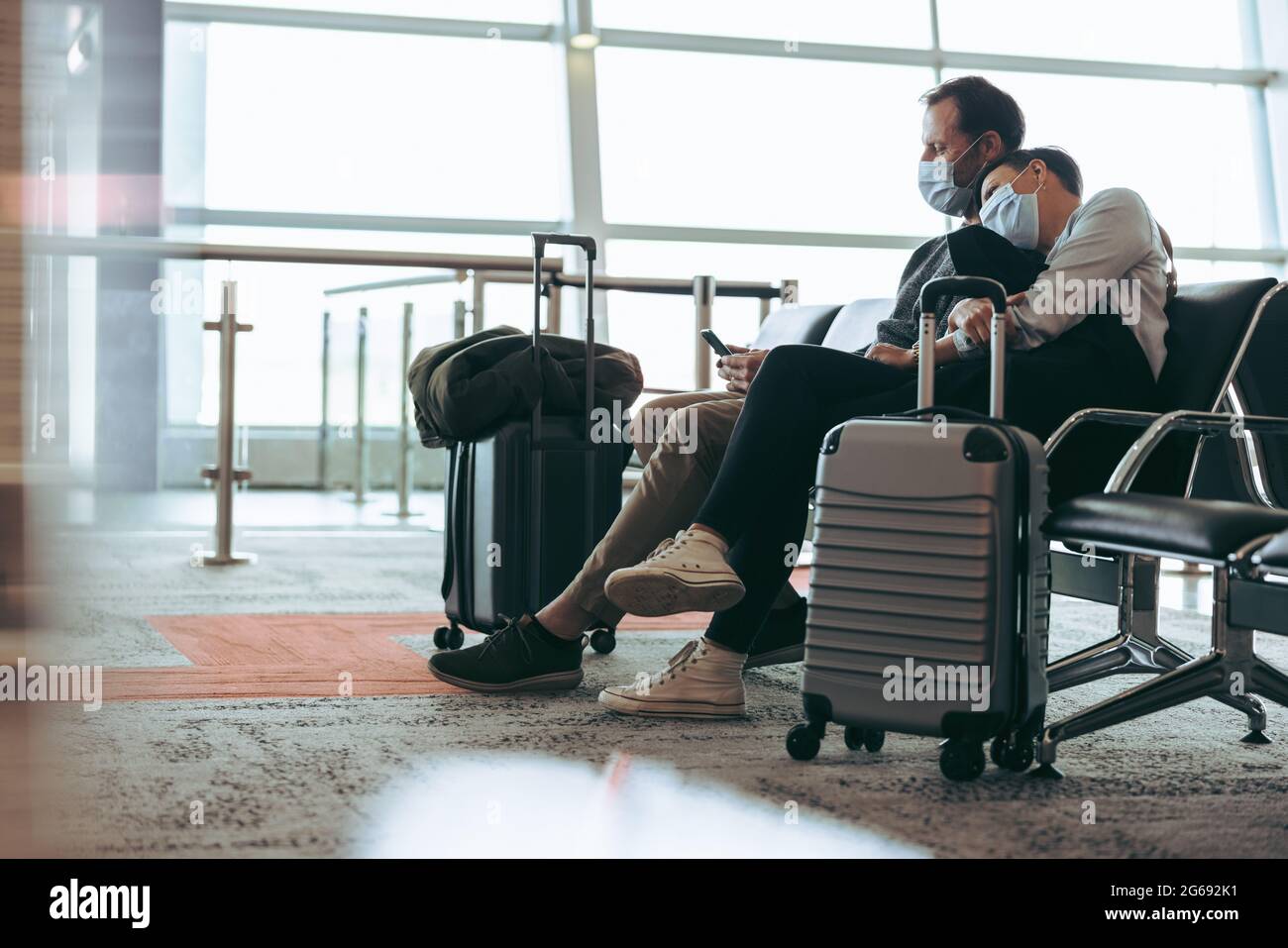 Ein Paar mit Gesichtsmasken sitzt am Flughafenterminal und wartet auf den Flug. Mann, der während der Korona neben einer Frau am Flughafen mit dem Handy sitzt Stockfoto