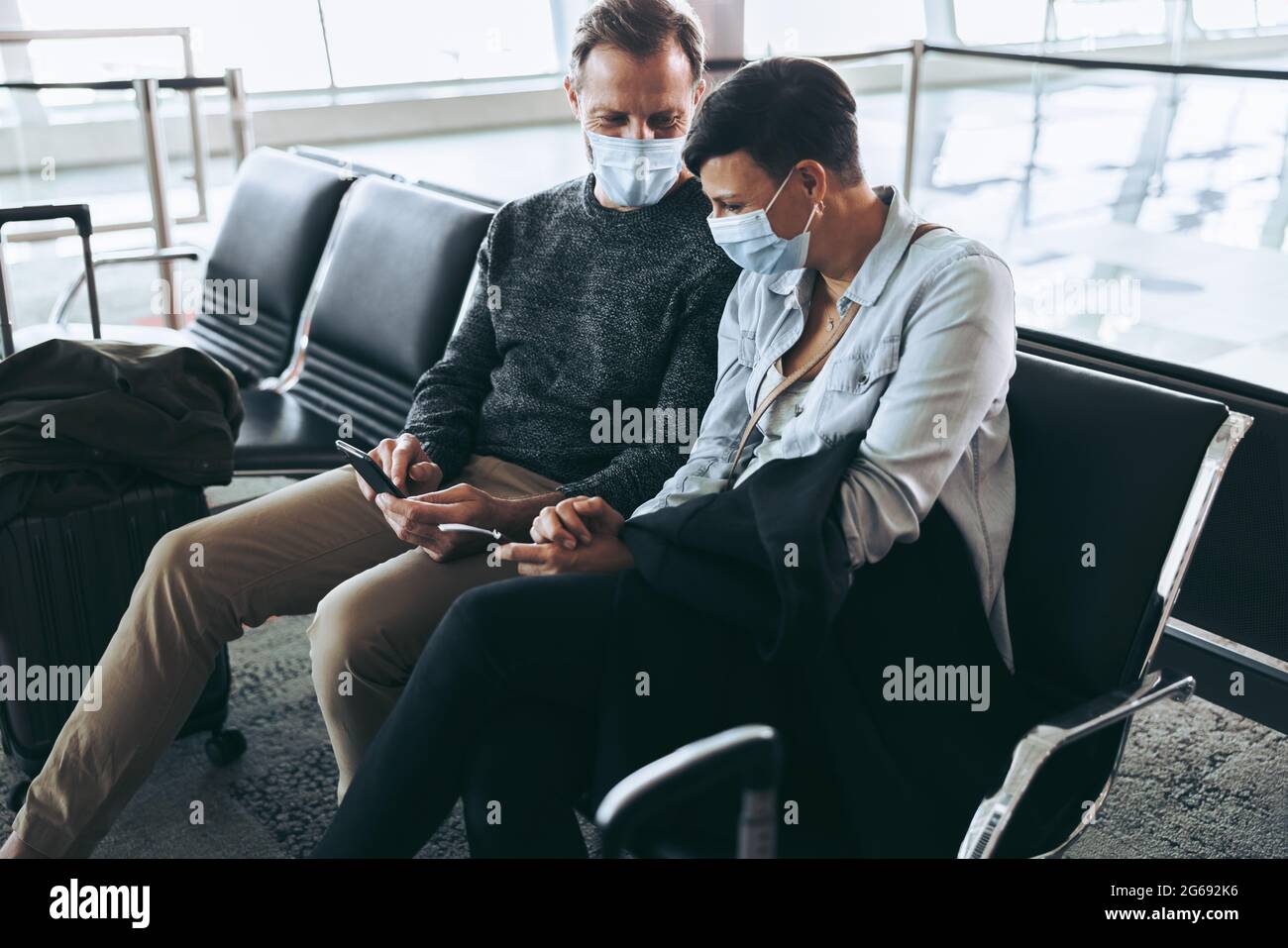 Paar mit Gesichtsmasken sitzen am Flughafen und mit dem Handy, um Fluginformationen zu überprüfen. Mann zeigt der Frau sein Handy, während er auf Sendung wartet Stockfoto