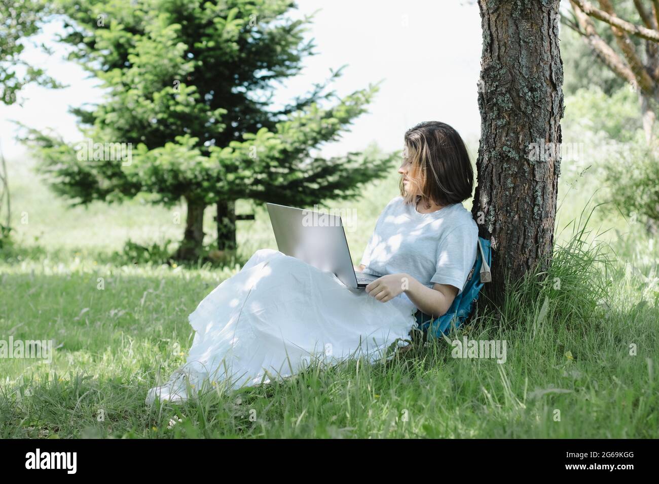 Nettes Teenager-Mädchen, das in der Nähe von Baum im Sommerpark mit offenem Laptop in den Händen sitzt und studiert. Konzept des Fernlernens Stockfoto