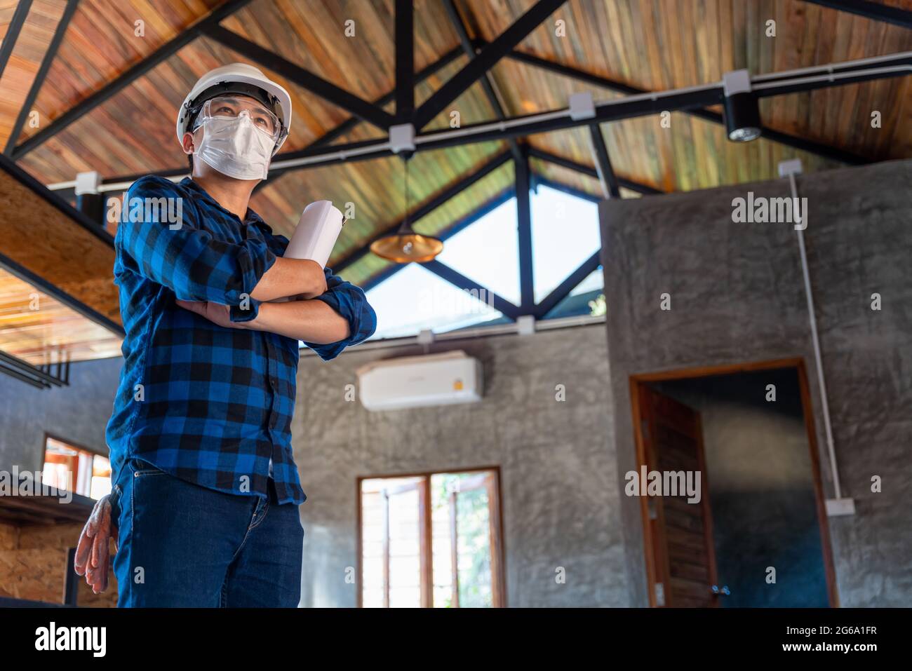 Glücklicher asiatischer Bauingenieur-Techniker, der mit gekreuzten Armen steht, nachdem er die Holzstruktur unter dem Dach bei Construction si überprüft hat Stockfoto