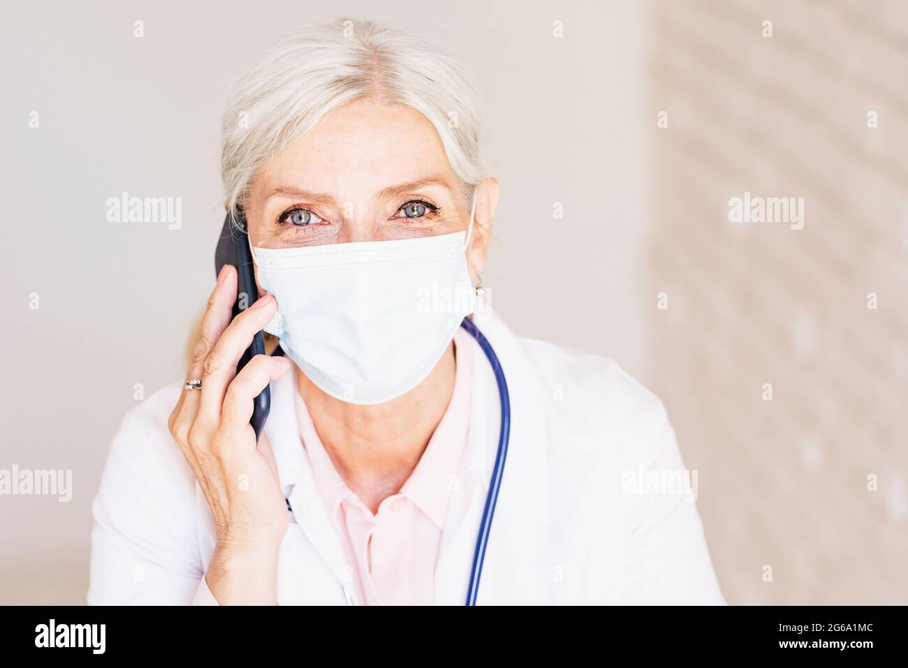 Aufnahme einer älteren Ärztin, die Gesichtsmaske trägt, während sie einen Anruf in der Arztpraxis hat. Stockfoto