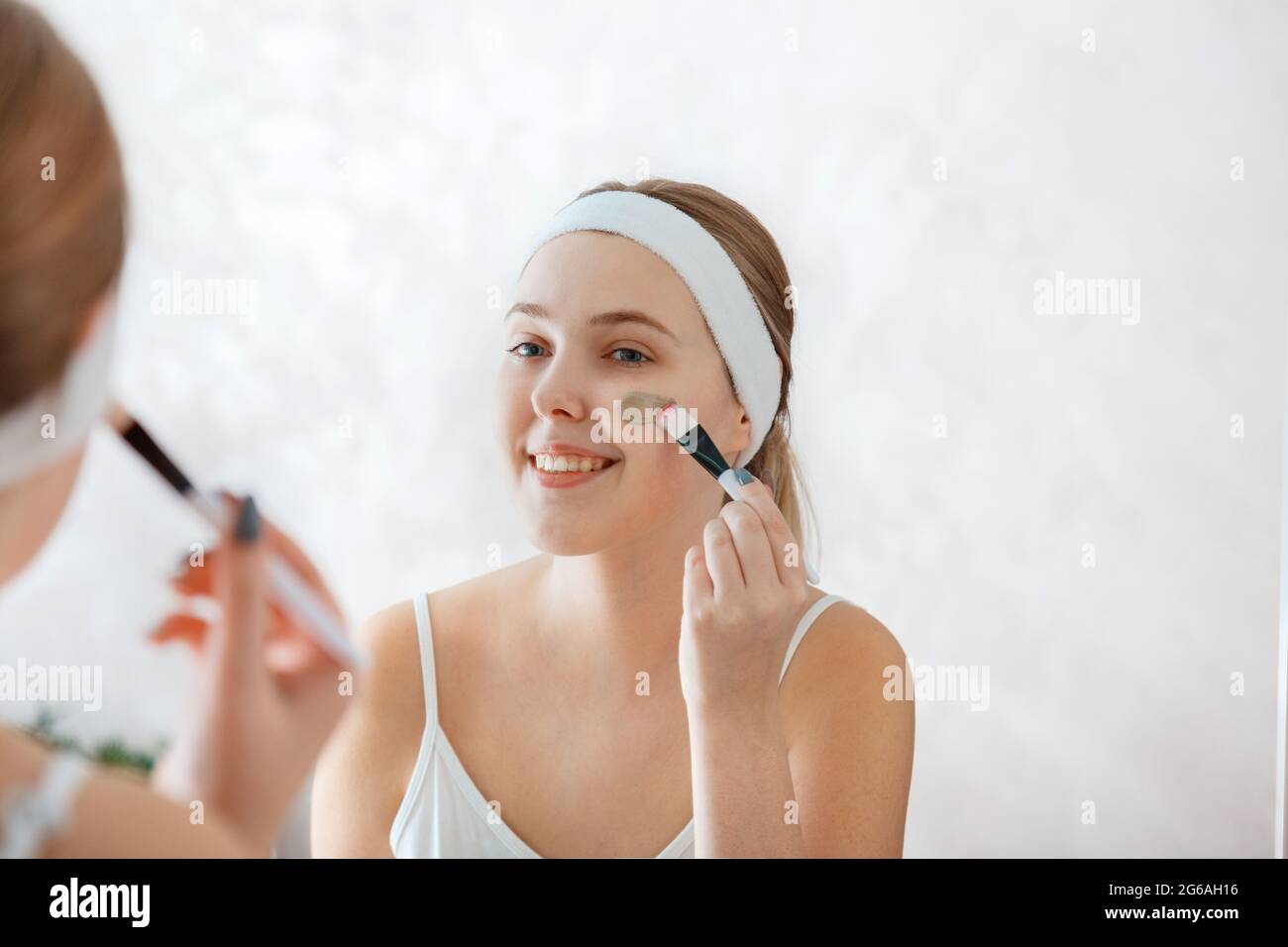 Junge Frau tragen Clay Gesichtsmaske. Junge Frau verwenden Hautpflege Schönheitsprodukte. Teenager-Mädchen breitet Tonmaske auf Wange mit Pinsel zu Hause im Badezimmer Stockfoto