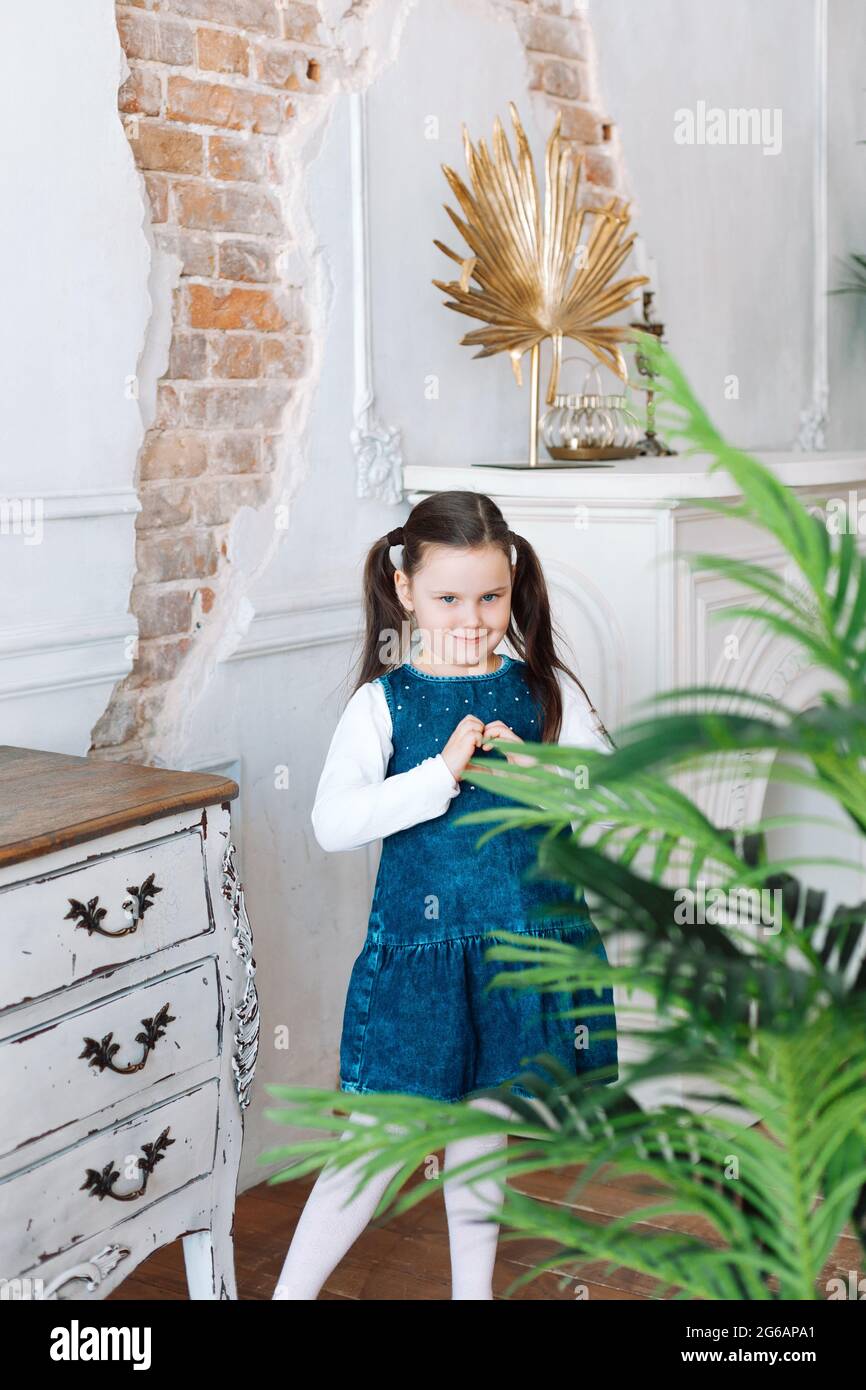 Ein Porträt einer schönen fünfjährigen Frau in einem Denim-Kleid, das mit den Fingern nach oben in einer häuslichen Umgebung mit Topflappen ein Herzsymbol macht Stockfoto