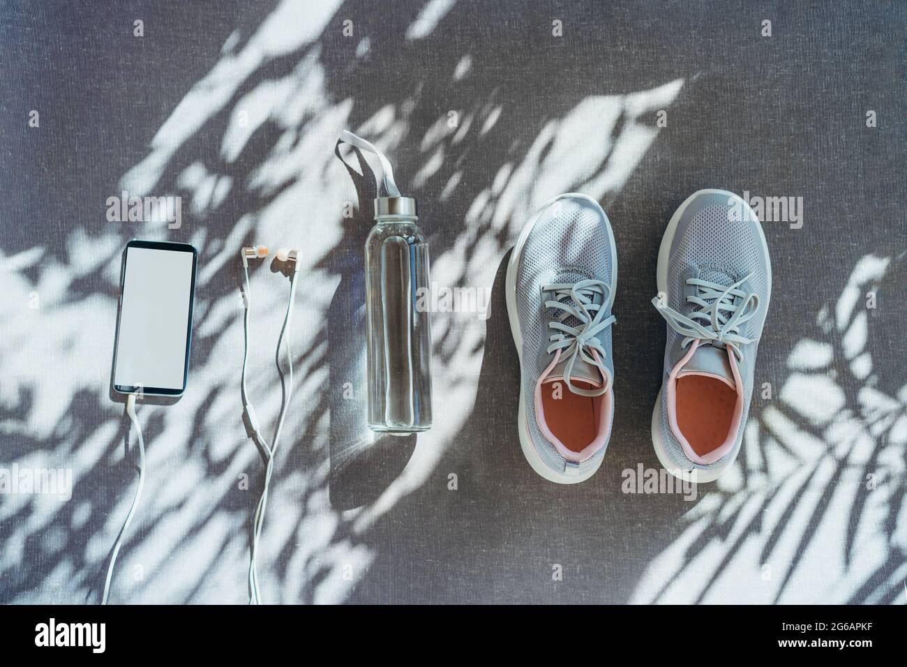 Draufsicht Flasche mit Wasser, leeres Smartphone, Kopfhörer und Sneaker auf grauem Hintergrund mit direktem Sonnenlicht und Schatten. Set für Laufen und Sport A Stockfoto