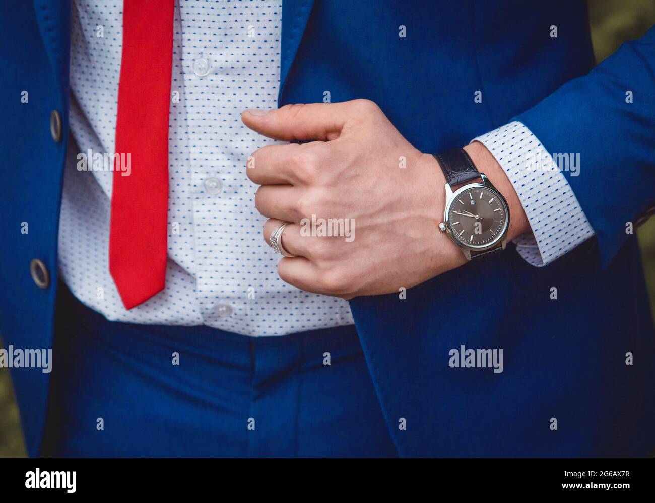 Die Hand eines Mannes mit einer Handuhr hält sich an einem blauen Anzug vor dem Hintergrund eines weißen Hemdes und einer roten Krawatte, einem Kleidungsstil für Geschäftsleute Stockfoto