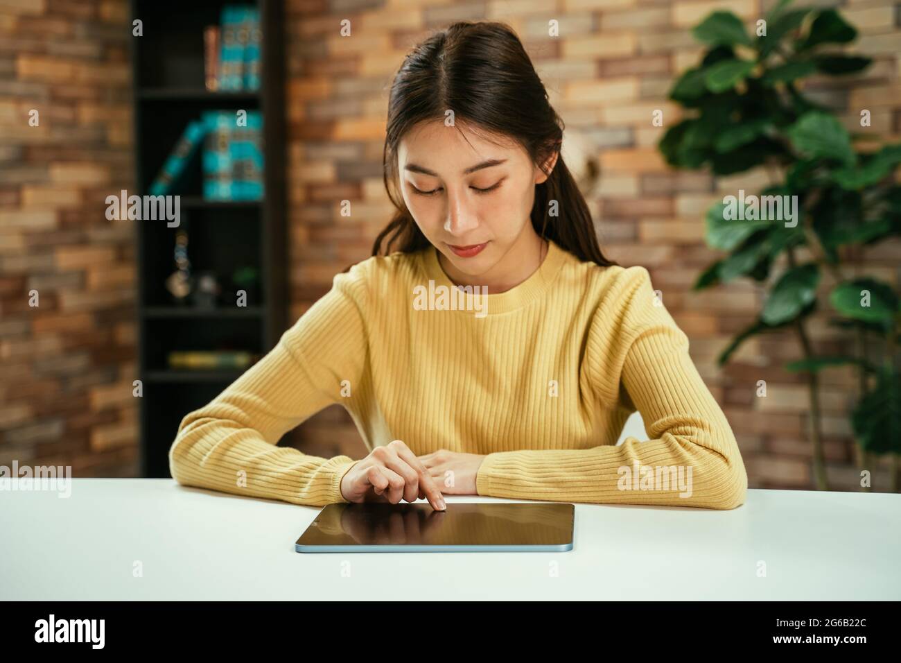 Fokussierte junge asiatische Frau, die zu Hause mit einem digitalen Tablet auf dem Schreibtisch sitzt, um in den sozialen Medien zu surfen und nachts zu Hause online zu shoppen Stockfoto