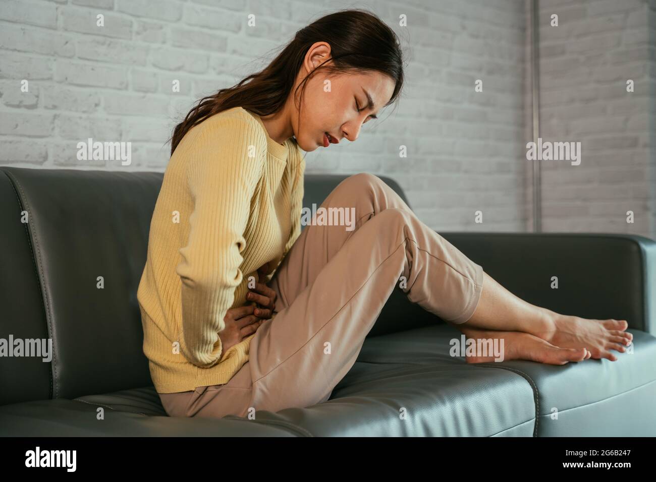 Traurige, verstörte und unglückliche junge asiatische Frau, die die Hände am Bauch hält und an Bauchschmerzen leidet, während die Augen zu Hause die Menstruation haben Stockfoto