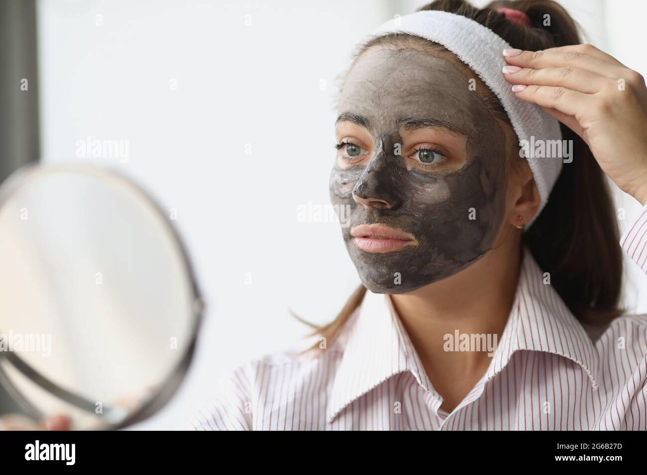 Junge Frau mit schwarzer Tonmaske, die zu Hause in den Spiegel schaut Stockfoto