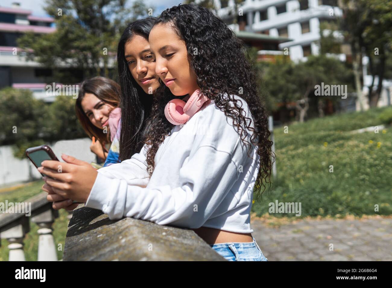 Ein lockiges Teenager-Mädchen und ihre Freundin mit brünettem Fell interagieren mit ihrem Handy, während ein anderes Mädchen sie in einem zweiten Einschuss unscharf sieht Stockfoto