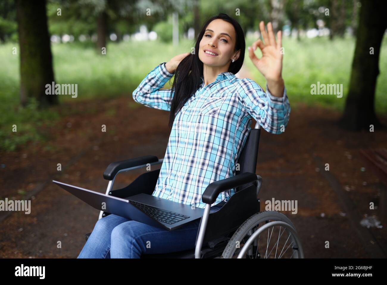 Eine behinderte Frau, die im Rollstuhl sitzt und einen Laptop auf ihrem Schoß hat und eine Geste zeigt, die in Ordnung ist Stockfoto
