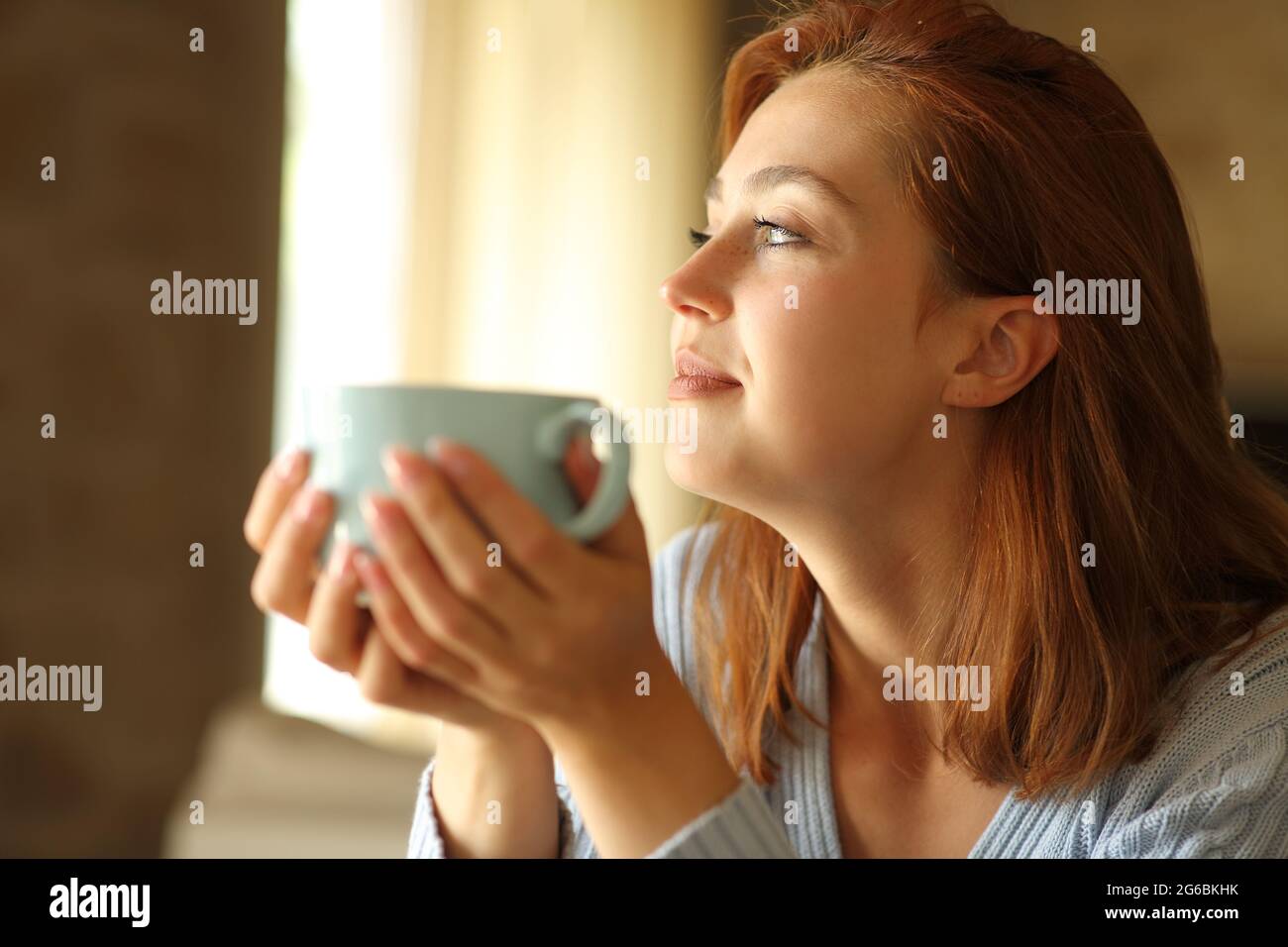 Zufriedene Frau, die zu Hause durch das Fenster Kaffee trinkt Stockfoto