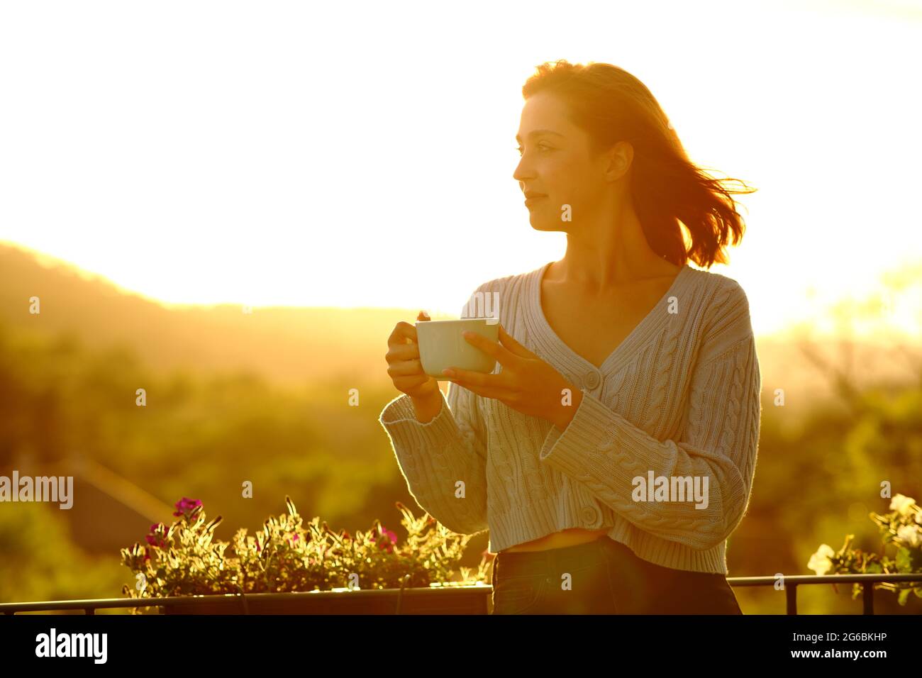Selbstbewusste Frau, die eine Kaffeetasse hält und auf einem Balkon den Sonnenuntergang betrachtet Stockfoto