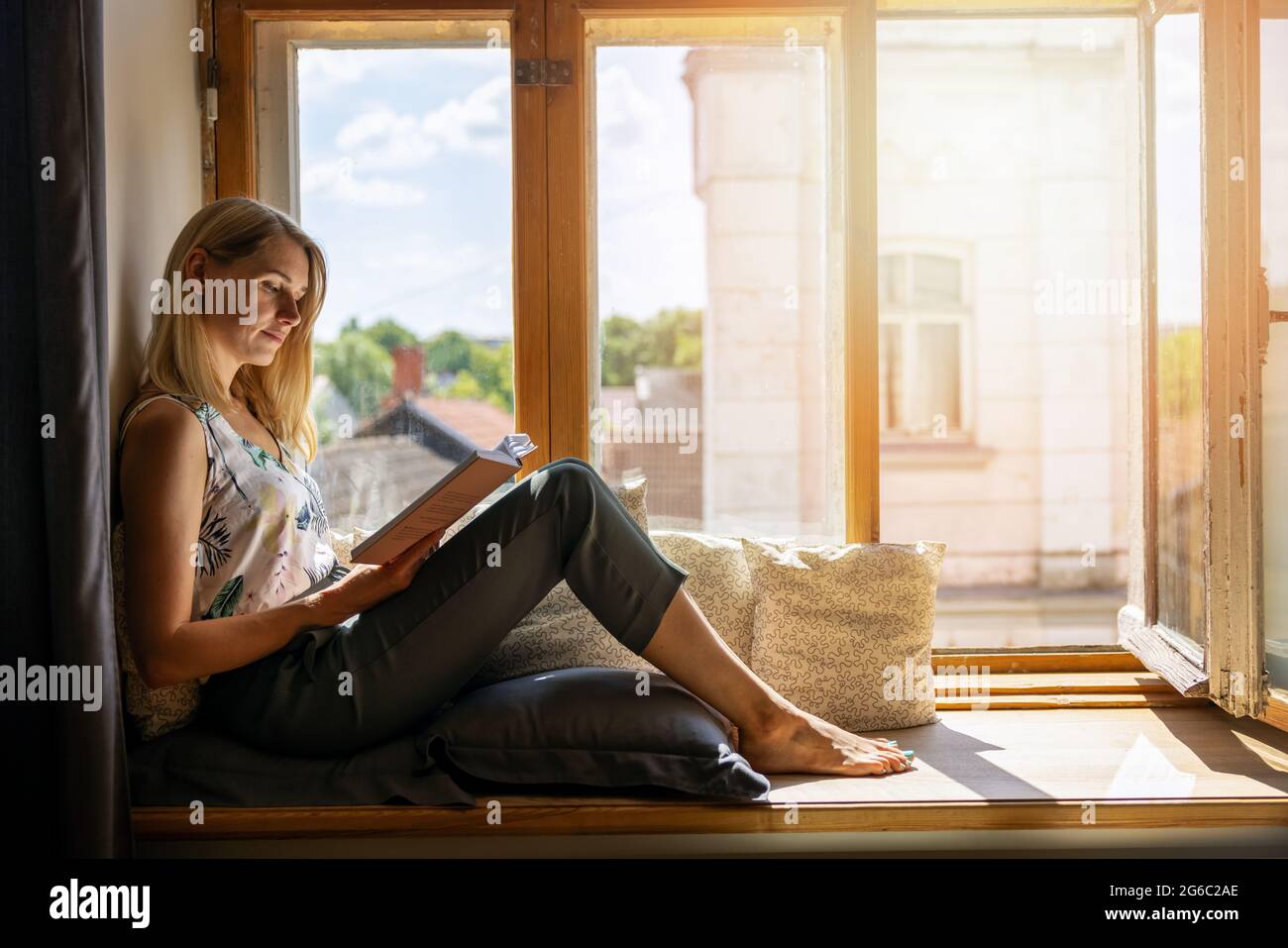 Junge Frau, die zu Hause auf einer gemütlichen, sonnigen Fensterbank sitzt und ein Buch liest Stockfoto