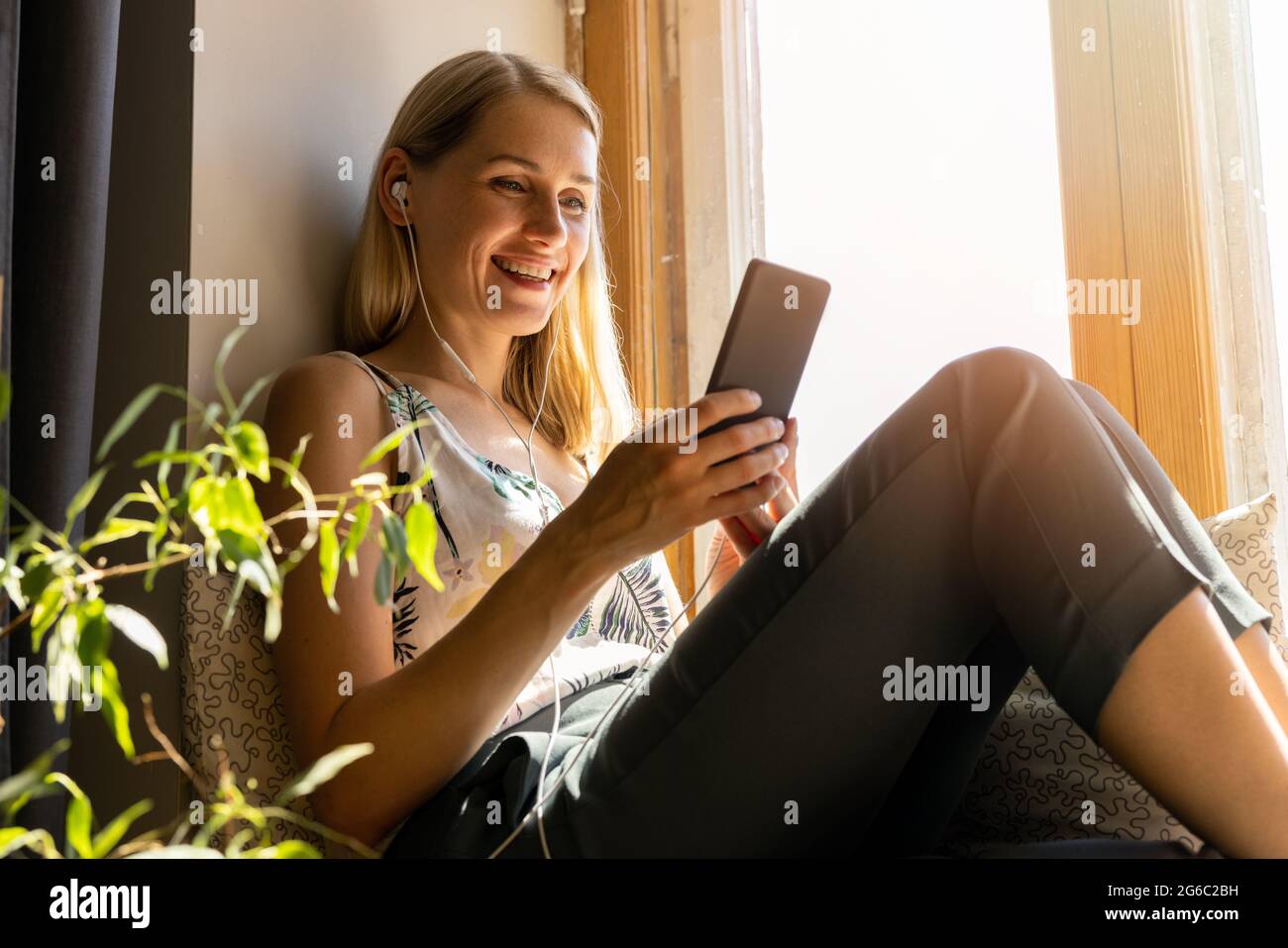 Lächelnde junge Frau, die auf der sonnigen Fensterbank sitzt und zu Hause das Smartphone benutzt Stockfoto