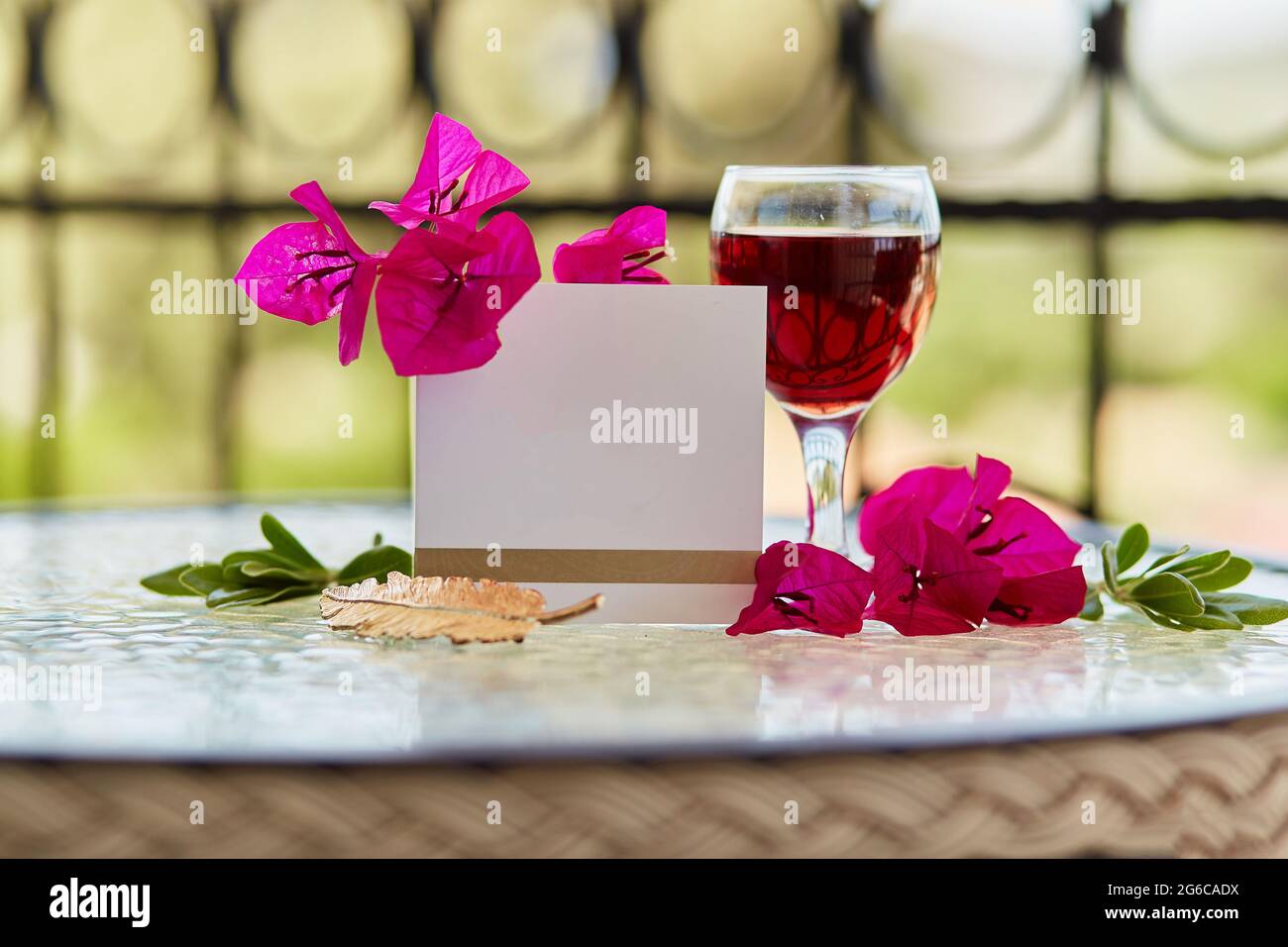Ein Glas Rotwein auf dem Tisch auf der Terrasse. Dekorative rosa Blüten von Bougainvillea und goldenen Federzeichen. Nachbau einer Postkarte oder Einladung. Romantisches Dinner-Konzept Stockfoto